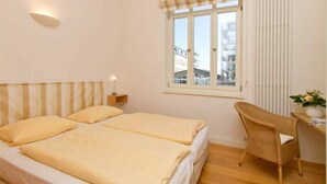 Ein Zimmer mit zwei Betten, einem Sessel, einem Fenster mit Jalousien, einer Wandleuchte und Blick auf Gebäude.