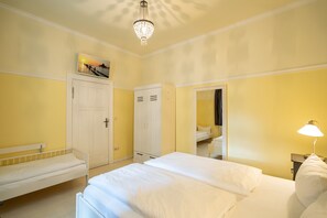 Quarto de hotel com cama, um banquinho, guarda-roupa, espelho e lustre.