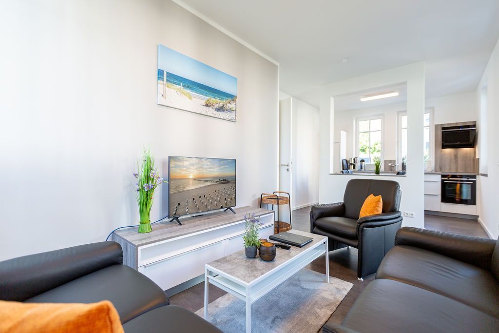 Ein modernes Wohnzimmer mit Flachbildfernseher, Couchtisch und Sofa. Im Hintergrund ist eine Küchenzeile mit Esstisch und Stühlen zu sehen.