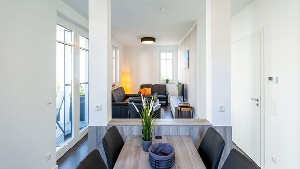 Een moderne eetruimte met een lange tafel, zwarte stoelen en een potplant. Achterin staat een bank en een televisie.