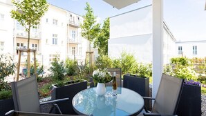 Een terras buiten met een ronde glazen tafel en vier stoelen, omringd door groen en gebouwen op de achtergrond.