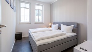 Een slaapkamer met een grijs bed, twee ramen, een nachtkastje met een lamp en een afstandsbediening op een witte bijzettafel.