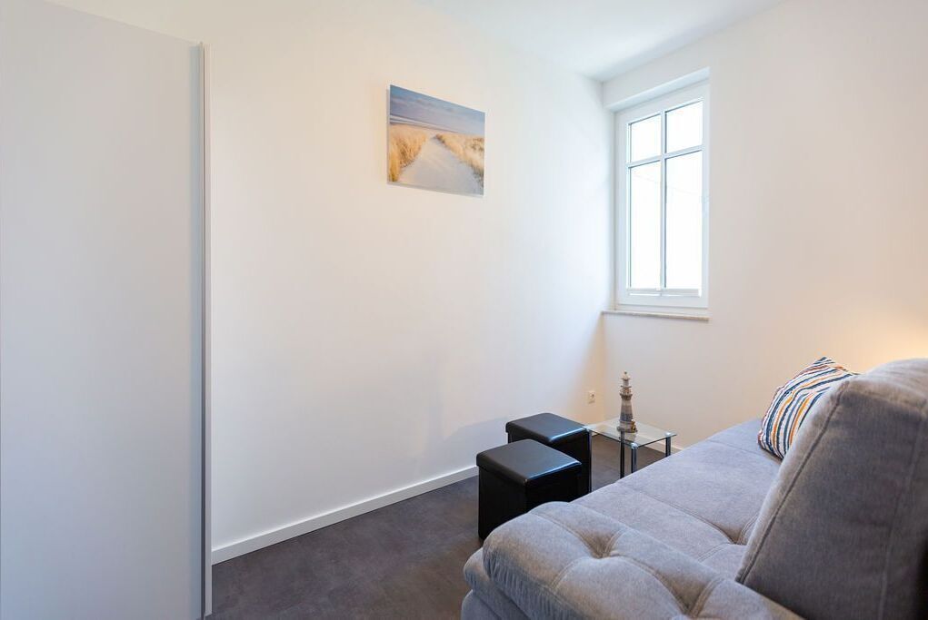 A room with a grey sofa, a small table with a lamp, a picture on the wall, and a window.