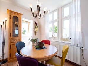 Un comedor con una mesa redonda de madera, cuatro sillas, una araña, un aparador con puertas de cristal y ventanas grandes con cortinas.