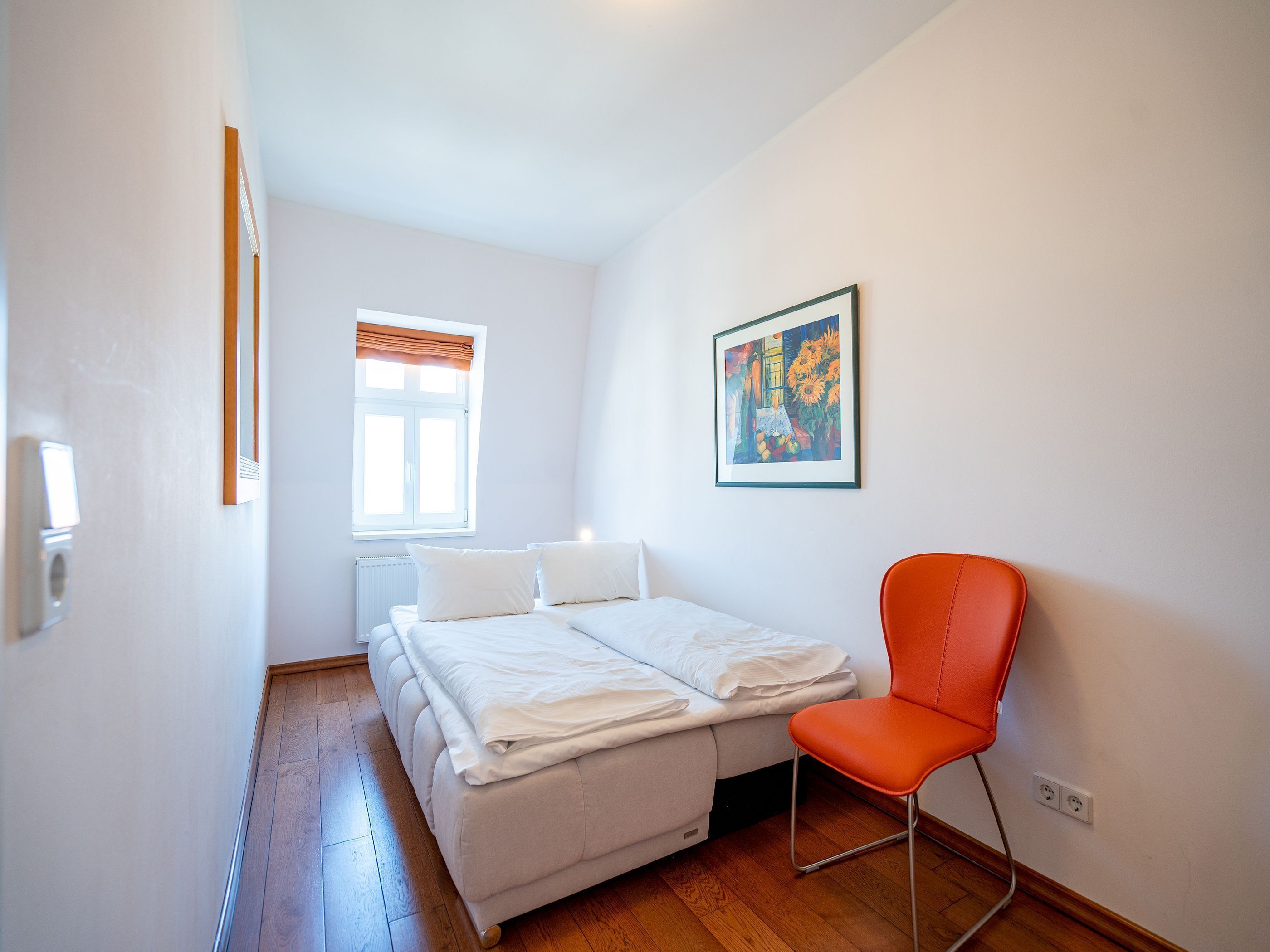 A bedroom with a bed, a chair, a window, a radiator, and a painting on the wall.