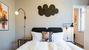 Ein ordentlich bezogenes Bett mit weißen Leinen und zwei Kissen, ein an der Wand befestigtes abstraktes Kunstwerk und ein Nachttisch mit einer Lampe.