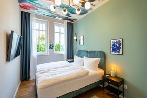 Un dormitorio con cama, una mesita de noche con lámpara, un televisor montado en la pared y una ventana con cortinas.