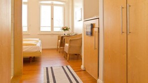 Een smalle gang met een bed, een zithoek met rieten stoelen en een houten garderobe.