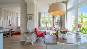 A modern living room with a wooden dining table, a red chair, and a beige sofa. There is a large window with curtains, a hanging lamp, and a vase with flowers on the table. - Rolandseck, App. 3, Germany (Heringsdorf)