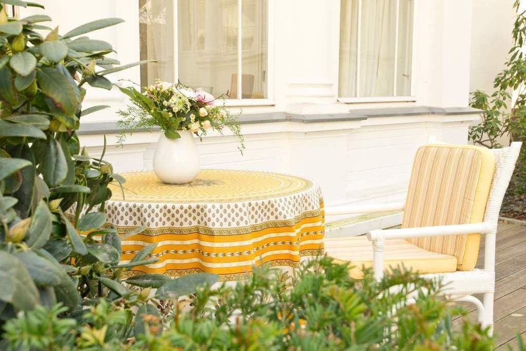 Una mesa redonda con mantel amarillo y blanco, un florero blanco con flores y una silla de mimbre con cojín amarillo.