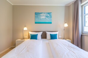 Uma cama de casal com roupa de cama branca, duas mesas de cabeceira com abajures, um quadro de um céu azul na parede e uma janela com cortinas.