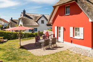 A red house with a thatched roof, a patio with a table and chairs, and a hedge in the foreground.