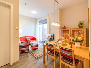 A modern living room with a red sofa, striped ottomans, a wooden dining table, and a TV in the background. - Quartier II, App. 2.1 (Heringsdorf)