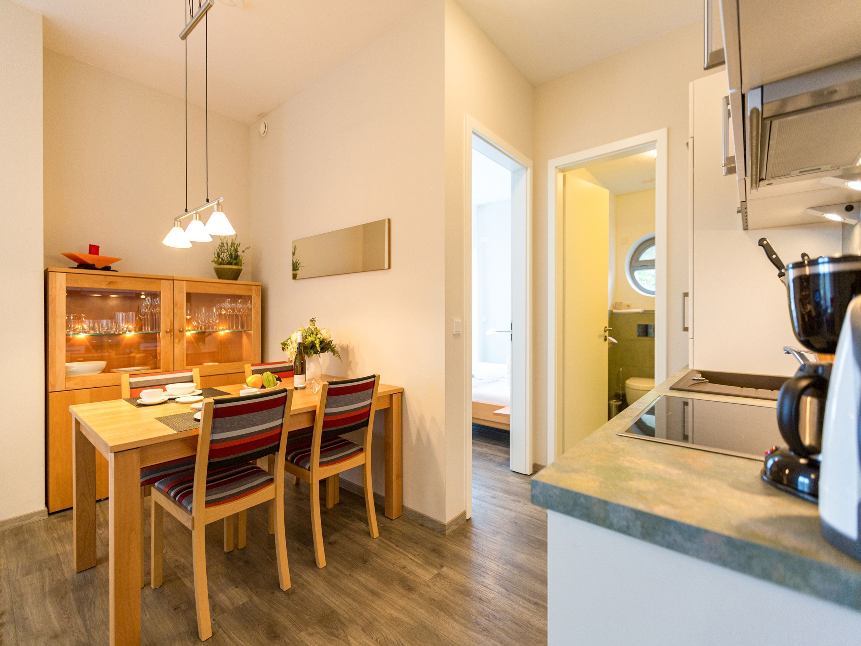 A modern kitchen with wooden cabinetry, a dining area with striped chairs, and a visible bathroom door.