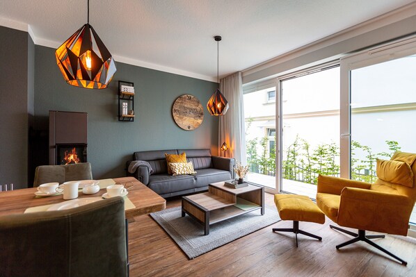 Una sala de estar moderna con sofá, mesa de centro y sillón amarillo. Hay una chimenea y un ventanal con vistas a zonas verdes.