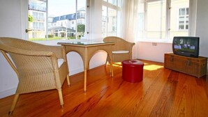 Una habitación con sillas de mimbre, una mesa de vidrio, un taburete rojo y un televisor sobre un soporte de madera.