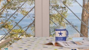 Uma mesa com toalha florida, uma caneca azul com estrela e vista para o mar através de uma janela com grade de metal.
