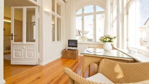 Una habitación luminosa con suelo de madera, un sofá de mimbre, una mesa de cristal y un amplio ventanal con cortinas.