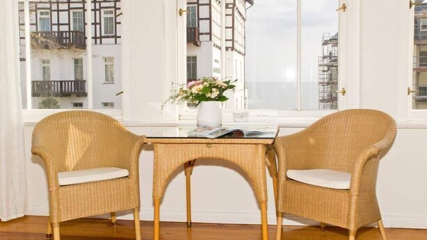 Una habitación con dos sillones de mimbre y una mesita, un jarrón con flores, y vistas a edificios a través de amplias ventanas.