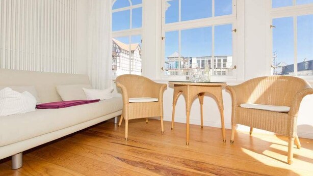 Ein Zimmer mit einer weißen Couch, zwei Korbstühlen, einem kleinen Tisch und großen, bogenförmigen Fenstern.