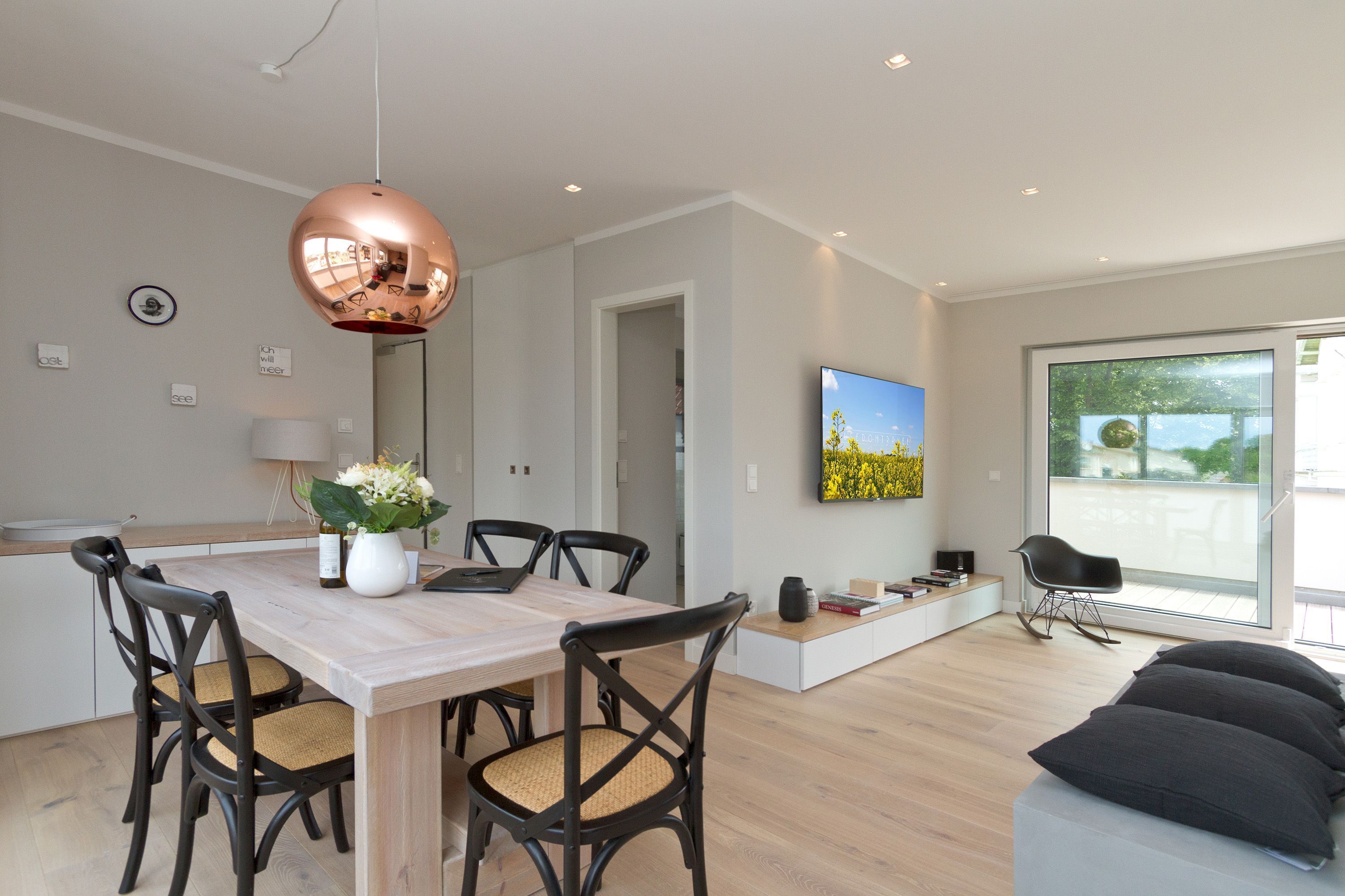 A modern living room with a dining area, featuring a wooden table, dark chairs, a large window, and a black sofa.