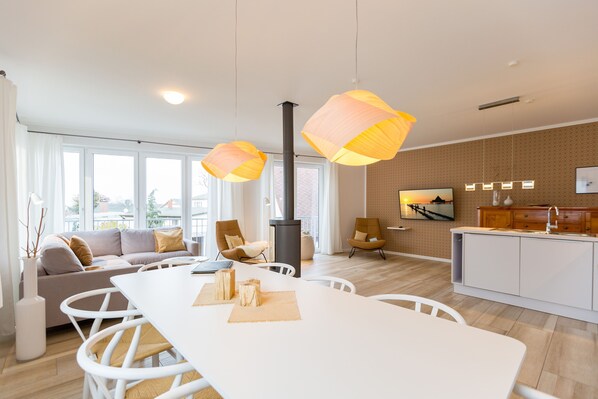 A modern living room with a white sofa, a dining table, and chairs. There is a kitchen area with a stove and cabinets. - Quartier Arinja, App. 01 (Heringsdorf)
