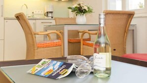 Un coin repas avec des chaises en osier, une table en verre, une bouteille de vin blanc et un magazine.