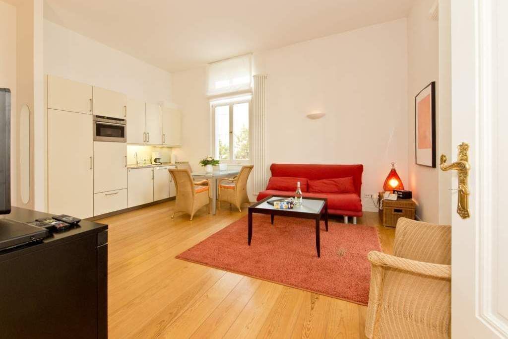 Een ruimtelijk ingerichte leefruimte met kitchenette, een rode bank, een salontafel en een eethoek met stoelen.