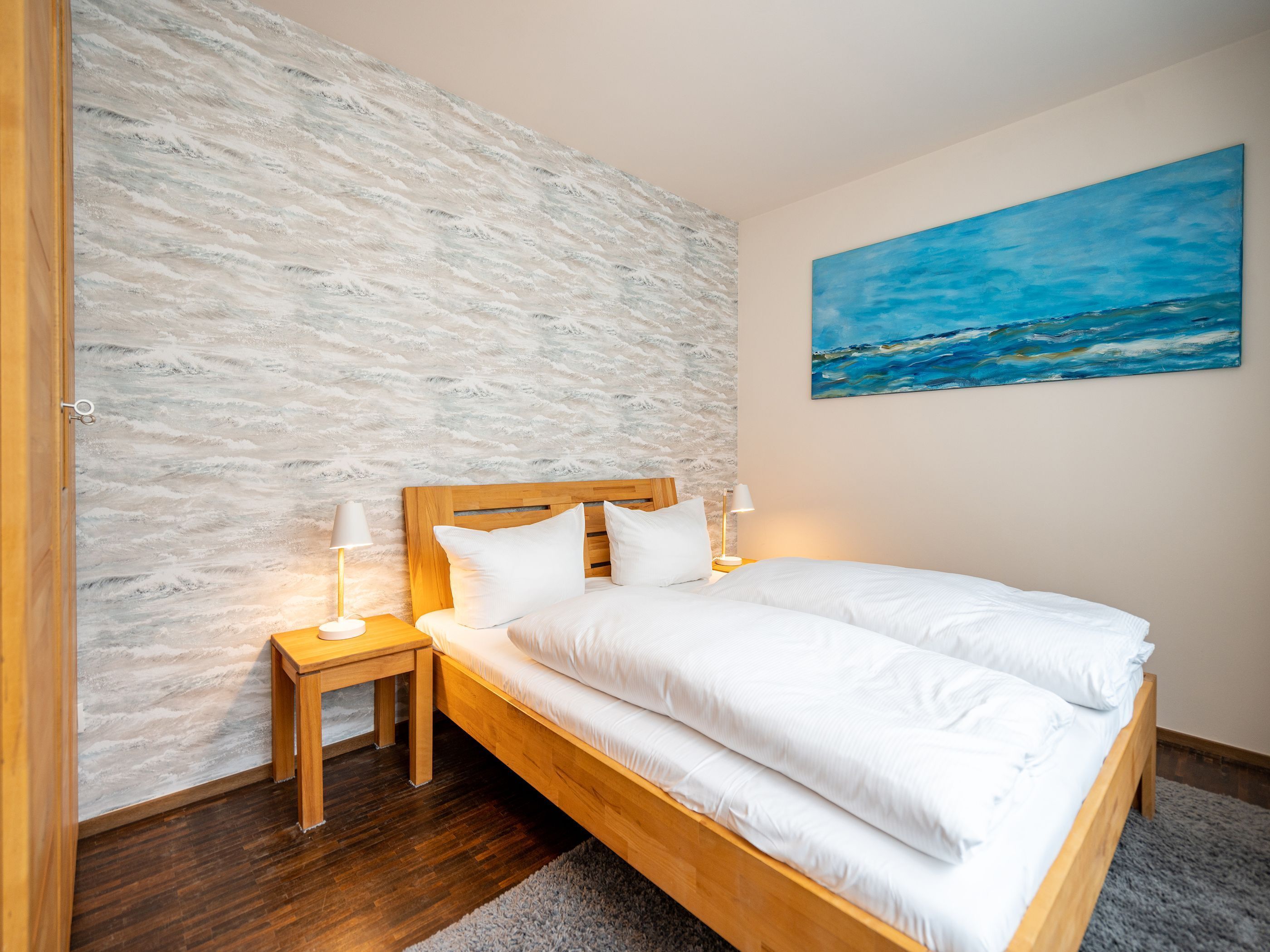 Een slaapkamer met een houten bed, wit beddengoed, een structuurwand, een schilderij en een nachtkastje met een lamp.