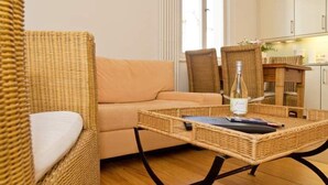 A wicker sofa and chairs in a room with a wooden floor, a small table with a bottle and a notebook, and a kitchen area in the background. - Villa Artemis, 01 Piraeus (Heringsdorf)