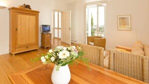 A room with a wooden dining table, a white vase with flowers, wicker furniture, a television, and a door leading outside. - Villa Artemis, 01 Piraeus (Heringsdorf)