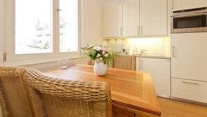 A modern kitchen with white cabinets, a wooden dining table, wicker chairs, and a vase with flowers. - Villa Artemis, 01 Piraeus (Heringsdorf)