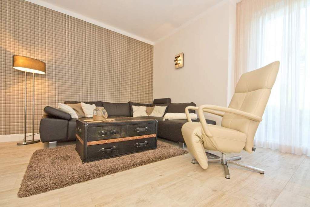 Un salon moderne avec un canapé d’angle, un fauteuil en cuir, une valise ancienne et un lampadaire.