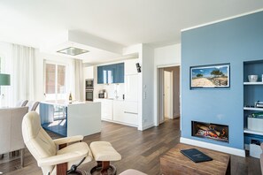 A modern living room with a fireplace, a dining area with a white table, and a kitchen with blue cabinets.