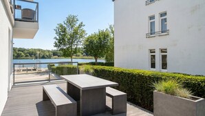 A modern outdoor patio with a table and benches, overlooking a lake and a white building with windows.