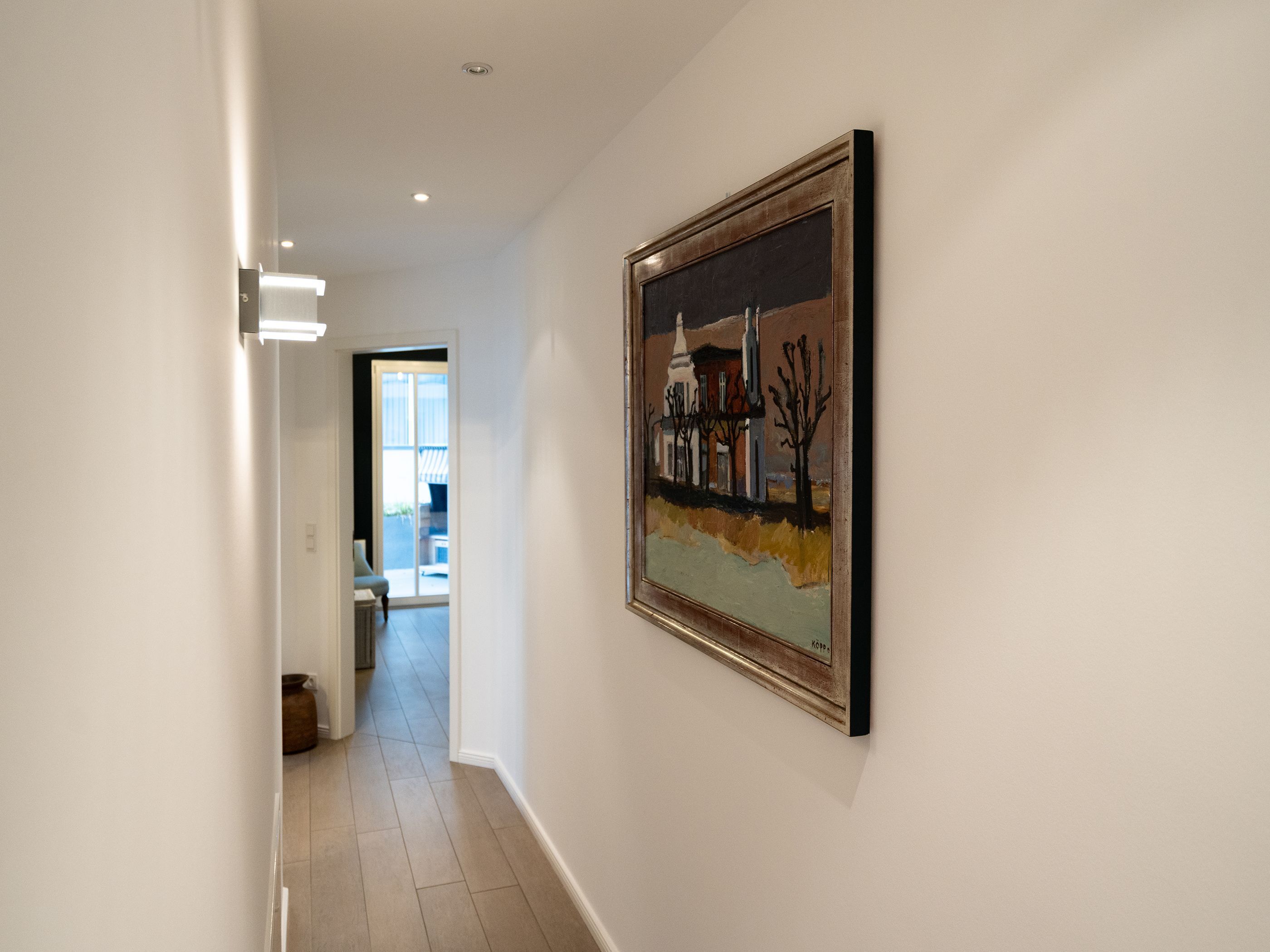 A hallway with a framed painting on the wall, a door leading to a room, and a view into another room.