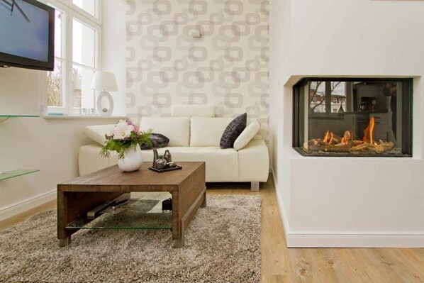 A living room with a white sofa, a glass coffee table, a fireplace, and a TV mounted on the wall.