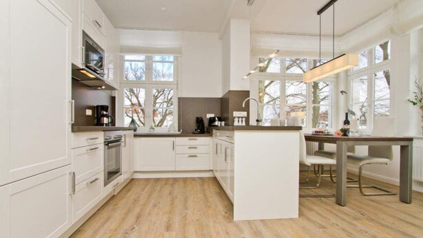 A modern kitchen with white cabinetry, a central island, and a dining area with a table and chairs. - Villa Waldschloss, app. 06 (Heringsdorf)