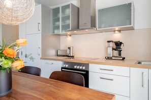 Una cocina moderna con armarios blancos, horno empotrado, cafetera y comedor con mesa y sillas de madera.