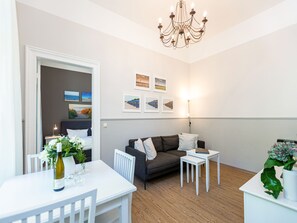 Ein modernes Wohnzimmer mit einem weißen Esstisch, einem dunkelgrauen Sofa und einer Chandelieber. An der Wand befinden sich gerahmte Bilder und auf dem Tisch steht ein Blumentopf.