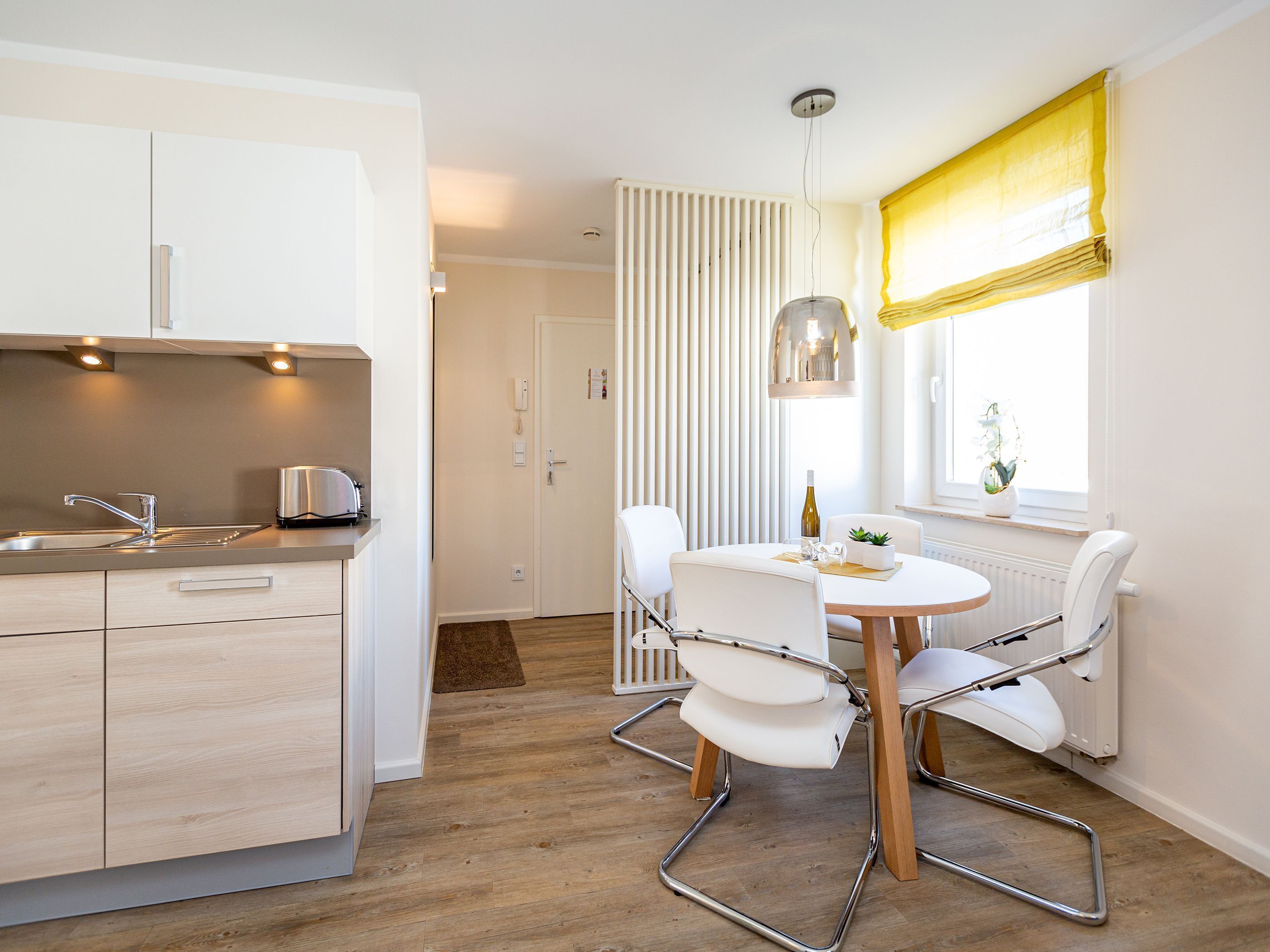 A modern kitchen with a sink, toaster, and white cabinets. There is a dining area with a round table, white chairs, and a pendant light.