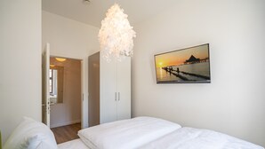 A bedroom with a bed, white bedding, a hanging light fixture, and a sunset painting on the wall. - Villa Waldschloss, app. 01 (Heringsdorf)
