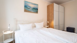 Una cama bien hecha con ropa blanca, una mesita de noche con lámpara, un cuadro colgado en la pared que representa una playa y un armario con puertas correderas.