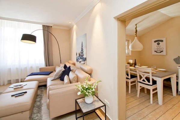 A modern living room with a beige sofa, a small side table with a vase, and a dining area with a white table and chairs.