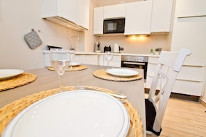 A dining table set with plates, cutlery, and wine glasses, surrounded by white chairs in a modern kitchen with white cabinets and a microwave.