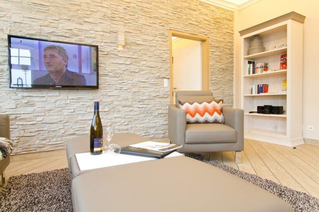 A modern living room with a stone wall, a television, a grey armchair with a patterned cushion, a bookshelf, and a bottle of wine on a table.