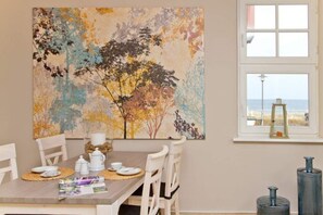 A dining area with a table set for four, a large abstract painting on the wall, and a window with a view of the beach.