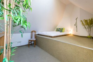 Quarto pequeno e compacto com cama, uma cadeira e mesa de cabeceira. Há plantas e uma prateleira na parede.