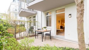 Een terras buiten met een tafel en stoelen, omgeven door groen en een gebouw-ingang.