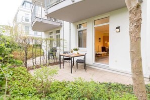 Eine Terrasse im Freien mit Tisch und Stühlen, umgeben von Grünpflanzen und einem Hauseingang.